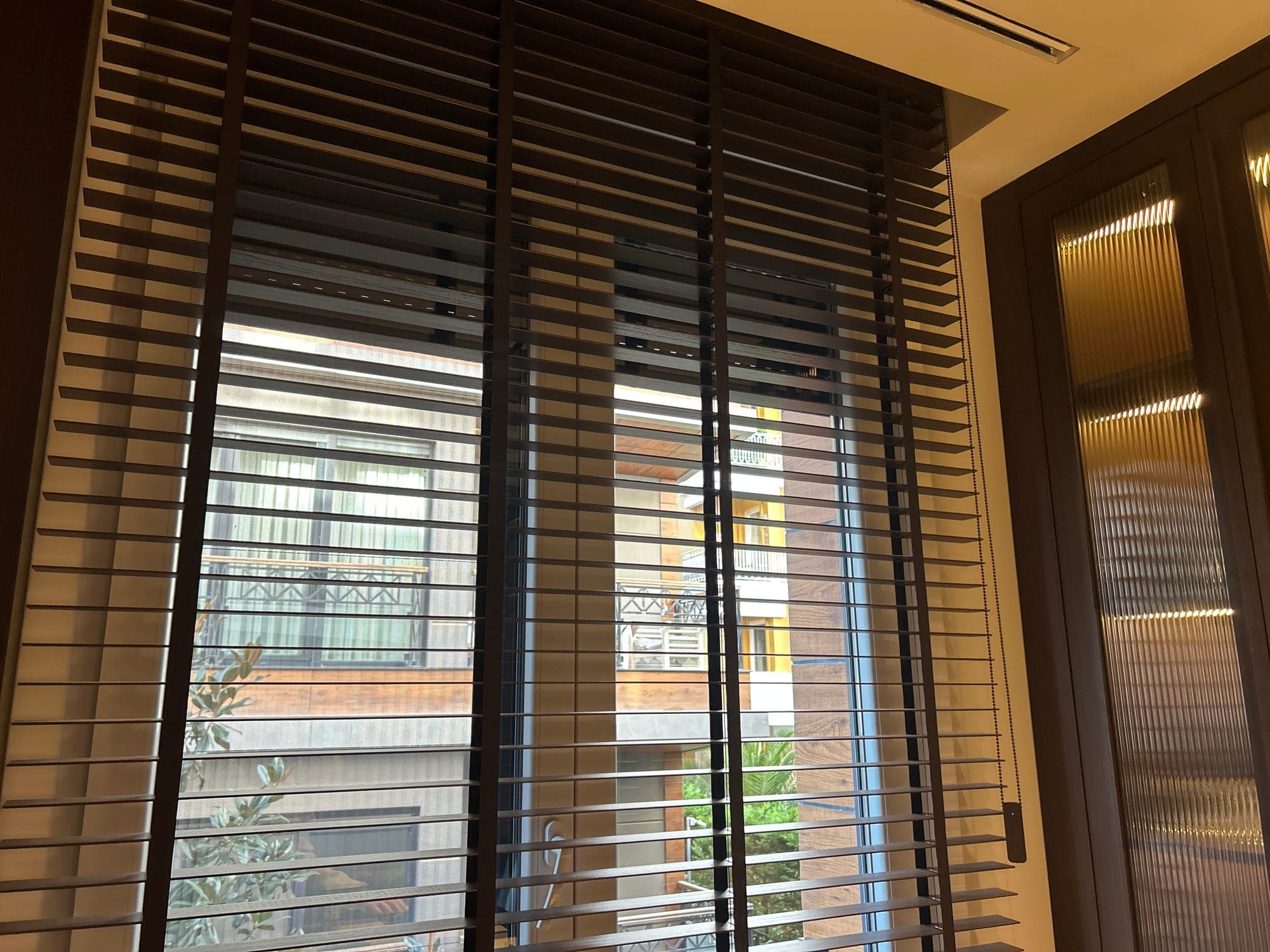 Open slats of a dark wooden Venetian blind with fabric tapes, beside a cabinet with fluted glass doors
