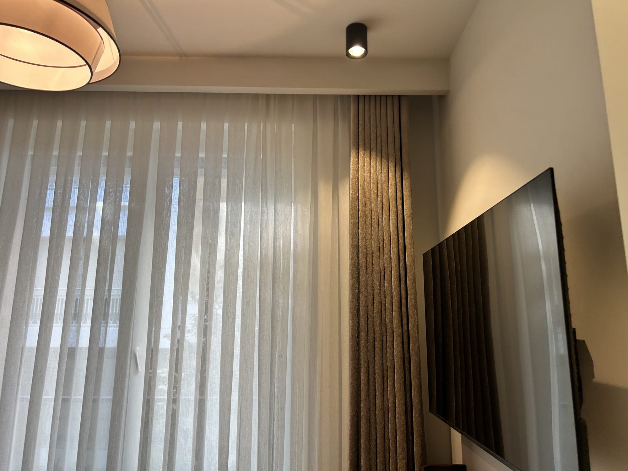 A room where the sheer and drapes fall from a recess in the ceiling, with a pendant lamp, spotlights and a television on the wall