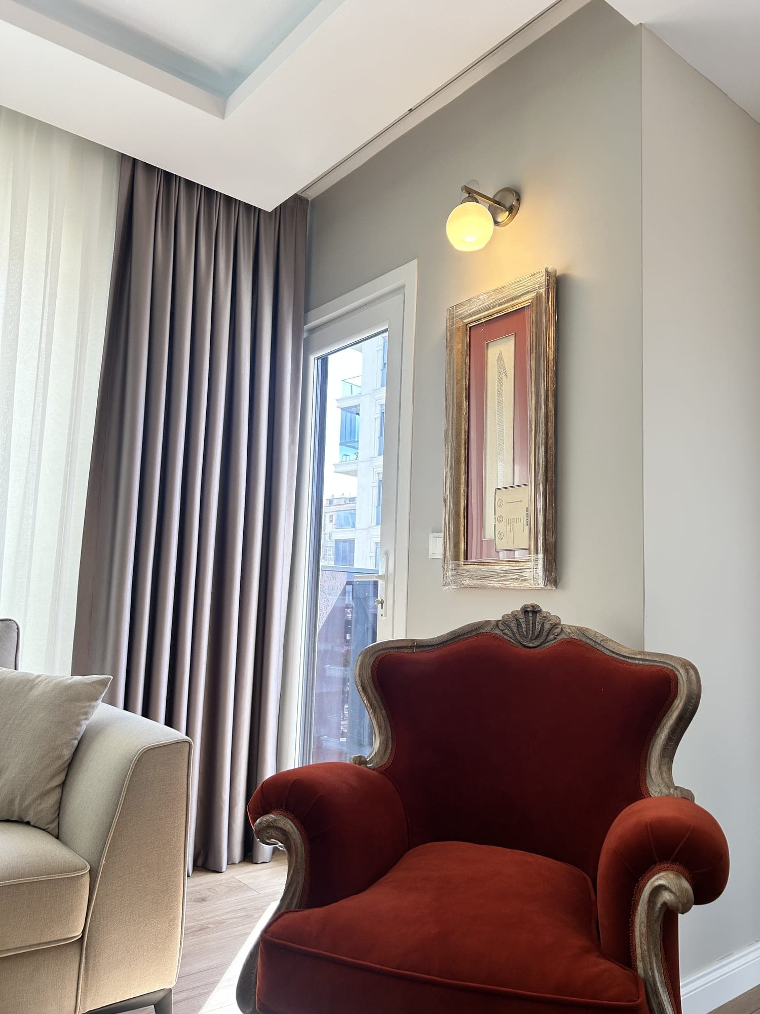 A grey drape and a sheer behind a red velvet armchair; a wall light and a framed picture on the wall
