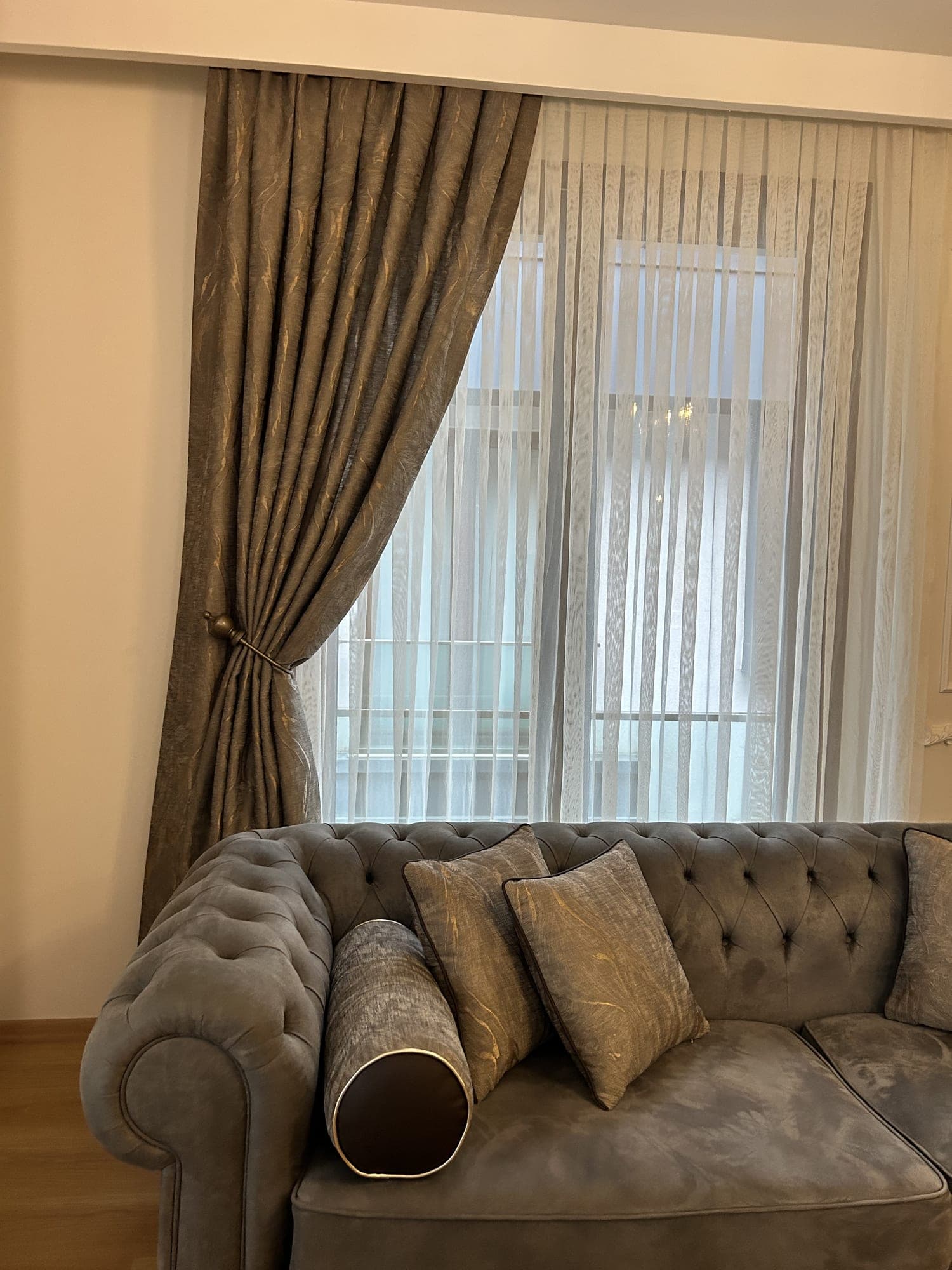 A patterned brown drape held aside by a holdback behind a Chesterfield sofa, and a sheer