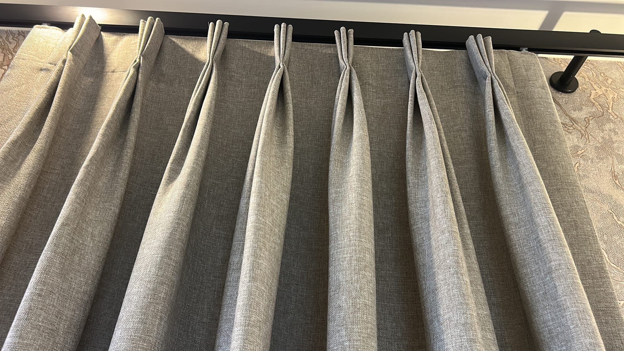The triple pinch pleats of a grey drape hung from a black pole; a patterned wall behind