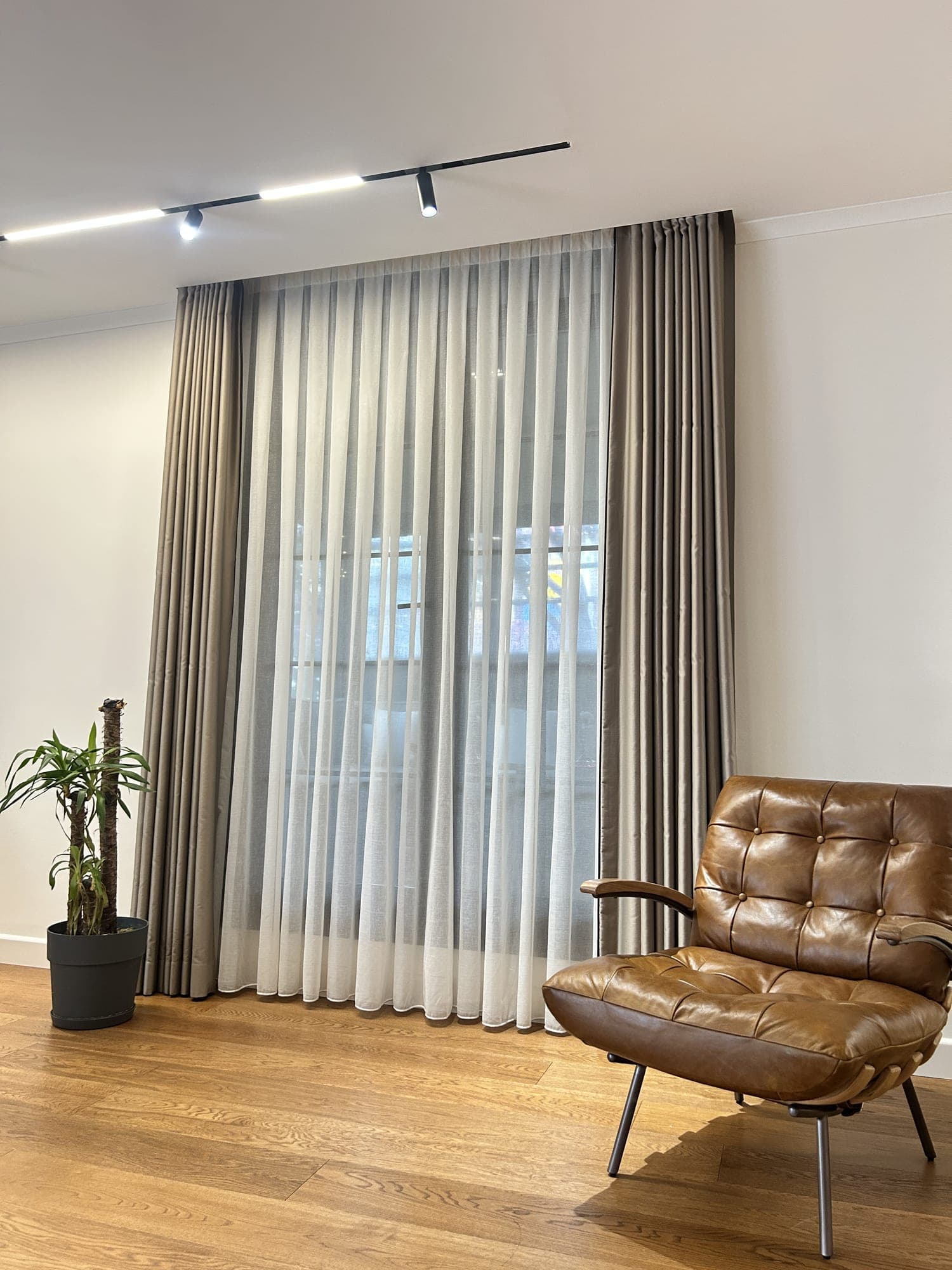 Taupe drapes falling to the floor from a track near the ceiling in a living room with spotlights, a sheer between them; a brown leather chair in front