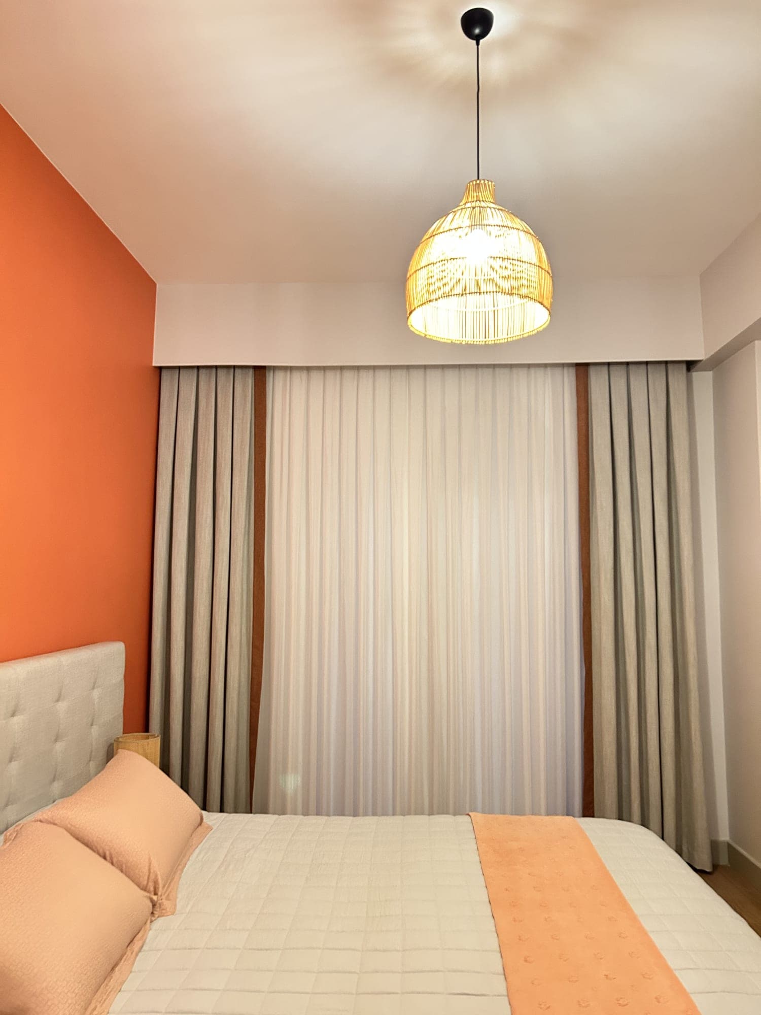 Drapes edged with a cinnamon band, and a sheer, in a bedroom with an orange wall; a woven pendant overhead